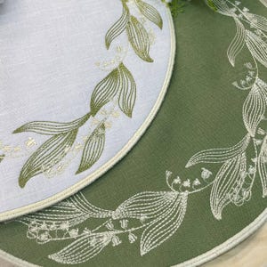May include: Two round placemats, one white and one green, with embroidered lily of the valley designs. The white placemat has a cream-colored border, while the green one has a matching green border. The embroidery features delicate green and white floral patterns.
