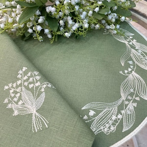 May include: A sage green linen napkin with white embroidered lily of the valley floral designs. A bouquet of white lily of the valley flowers rests on the napkin. The napkin has a white border.