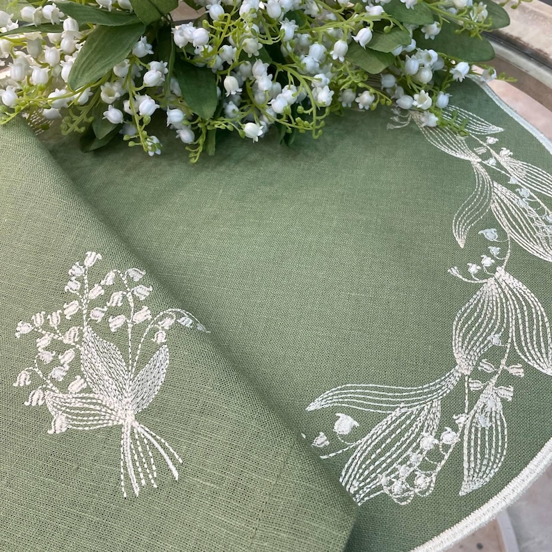 May include: A sage green linen napkin with white embroidered lily of the valley floral designs. A bouquet of white lily of the valley flowers rests on the napkin. The napkin has a white border.