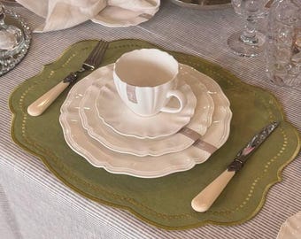 May include: A white teacup and saucer set with a scalloped edge, placed on a green placemat. A fork and knife with cream-colored handles are on the placemat. The table setting includes a folded napkin and glassware.