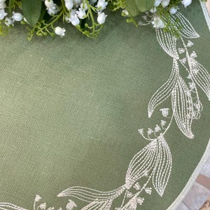 May include: A round, sage green linen tablecloth with white embroidered lily of the valley detailing along the edge. Fresh lily of the valley flowers are arranged at the top. The tablecloth is on a wooden table.
