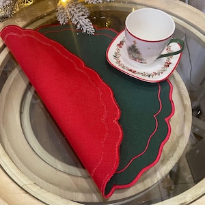 Può includere: Un'apparecchiatura da tavola a tema natalizio con un sottobicchiere rosso e verde smerlato. Una tazza e un piattino bianchi con un disegno di albero di Natale sono posizionati sul sottobicchiere. La tazza ha un manico verde e un bordo rosso.