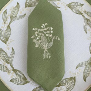 May include: A folded green linen napkin with an embroidered lily of the valley design. The napkin is placed on a white placemat with a matching embroidered floral border. The embroidery is in a light beige color.