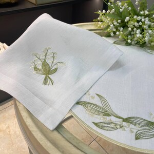May include: White linen napkin and table runner set with embroidered lily of the valley floral design in green and cream. The napkin is square, and the table runner has a scalloped edge.