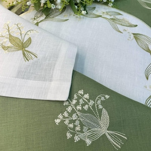 May include: White and green linen napkins and placemats with embroidered lily of the valley floral designs. The napkins are folded, and the placemats are partially visible. The embroidery features shades of green and white.