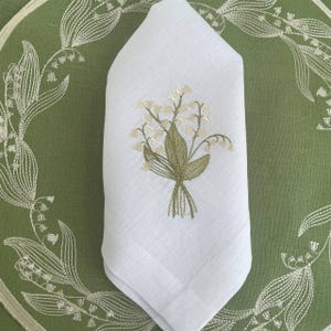 May include: White linen napkin with an embroidered bouquet of lily of the valley flowers. The napkin is folded and placed on a green tablecloth with a lily of the valley design.