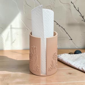 May include: A cylindrical peach-coloured paper towel holder featuring a vertical cutout and embossed floral designs including tulips and daisies on the exterior surface, holding a roll of white paper towels with a textured geometric pattern.