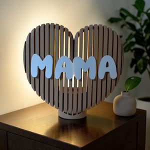 May include: A heart-shaped wooden lamp with the word "MAMA" in white letters. The lamp has a light beige color and is made of vertical wooden slats. It sits on a dark brown wooden surface.