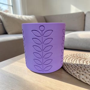 May include: A cylindrical, lavender-colored planter with a repeating leaf pattern. The planter is made of a solid material and sits on a wooden surface. The design is a modern take on a classic botanical theme.