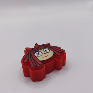 Peut inclure: Un emporte-pi&egrave;ce en plastique rouge en forme de t&ecirc;te de personnage de dessin anim&eacute; avec de longs cheveux rouges, un visage cr&egrave;me, un petit sourire et des yeux repr&eacute;sent&eacute;s par une croix noire et un cercle noir, avec un cadre &eacute;pais et solide.