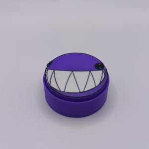 Peut inclure: Un r&eacute;cipient rond violet dot&eacute; d'un couvercle orn&eacute; d'un visage souriant. Le visage pr&eacute;sente deux petits yeux noirs avec des reflets bleus et une large bouche remplie de grandes dents triangulaires blanches, soulign&eacute;es de noir sur fond violet.