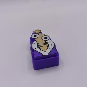 Peut inclure: Un charm rectangulaire violet en forme de bloc repr&eacute;sentant une pi&egrave;ce de roi d'&eacute;checs beige portant une cape blanche et noire style hermine. La pi&egrave;ce de roi a deux grands yeux blancs avec des pupilles noires sur une base violette.