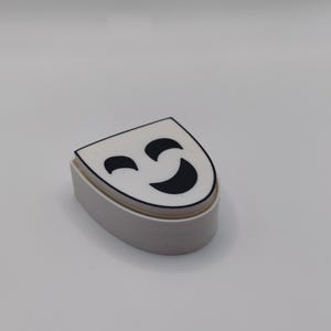 Peut inclure: Un objet blanc en forme de demi-cercle avec une surface sup&eacute;rieure plate pr&eacute;sentant un graphique noir repr&eacute;sentant un visage qui fait un clin d'&oelig;il. Le visage a une ligne courbe pour un &oelig;il ferm&eacute; et une forme courbe plus grande pour une bouche souriante.