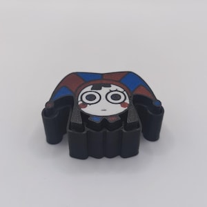 Peut inclure: Un emporte-pi&egrave;ce en plastique noir repr&eacute;sentant un personnage inspir&eacute; d'un bouffon avec un visage blanc, de grands yeux circulaires et un chapeau rouge et bleu. Le personnage a des marques rouges sur les joues et une bouche en trait simple.