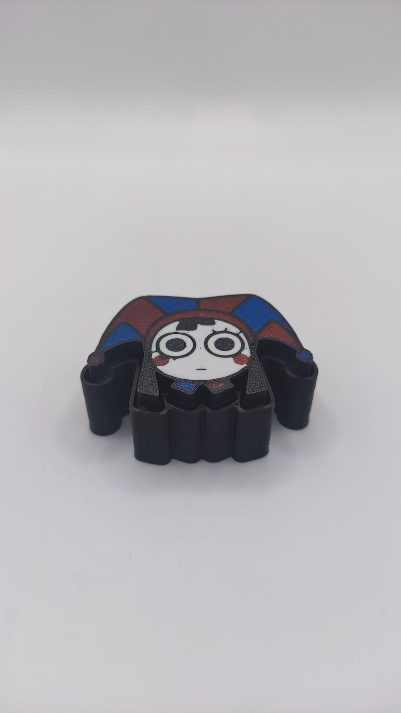 Peut inclure: Un emporte-pi&egrave;ce en plastique noir repr&eacute;sentant un personnage inspir&eacute; d'un bouffon avec un visage blanc, de grands yeux circulaires et un chapeau rouge et bleu. Le personnage a des marques rouges sur les joues et une bouche en trait simple.