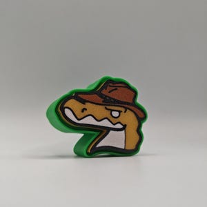 Peut inclure: Un emporte-pi&egrave;ce en plastique vert en forme de t&ecirc;te de dinosaure portant un chapeau fedora marron. Le dinosaure a un visage beige, une m&acirc;choire blanche et un contour noir. Le design pr&eacute;sente une esth&eacute;tique de dessin anim&eacute; stylis&eacute;e.