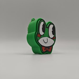 Peut inclure: Un porte-cl&eacute;s vert vif en forme de grenouille repr&eacute;sentant un visage de dessin anim&eacute; avec de grands yeux blancs, des pupilles noires, des joues rouges et un n&oelig;ud papillon rouge. Le personnage a un museau blanc et une bouche en trait noir.