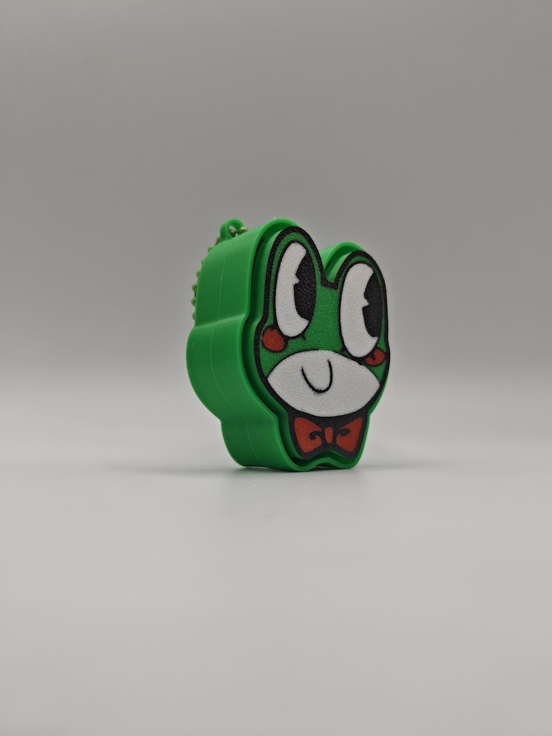 Peut inclure: Un porte-cl&eacute;s vert vif en forme de grenouille repr&eacute;sentant un visage de dessin anim&eacute; avec de grands yeux blancs, des pupilles noires, des joues rouges et un n&oelig;ud papillon rouge. Le personnage a un museau blanc et une bouche en trait noir.