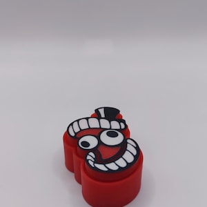 Peut inclure: Un emporte-pi&egrave;ce rouge en forme de t&ecirc;te de personnage de dessin anim&eacute; avec un large sourire &eacute;dent&eacute;. La surface sup&eacute;rieure pr&eacute;sente un motif graphique noir et blanc repr&eacute;sentant de grands yeux, un petit chapeau noir et une bouche aux dents carr&eacute;es.
