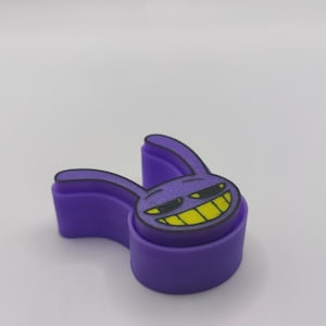 Peut inclure: Un emporte-pi&egrave;ce en plastique violet en forme de t&ecirc;te de lapin de dessin anim&eacute; avec de longues oreilles. La surface sup&eacute;rieure pr&eacute;sente un graphique imprim&eacute; d'un visage de lapin avec des yeux pliss&eacute;s et un large sourire aux dents jaunes et noires.