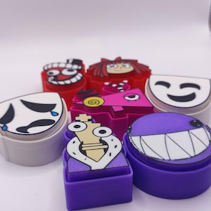 Op de afbeelding: Een verzameling kleurrijke stempels met cartoongezichten in verschillende vormen. De stempels hebben ontwerpen met rode, paarse, roze, witte en grijze kleuren. De stempels zijn gerangschikt op een wit oppervlak.