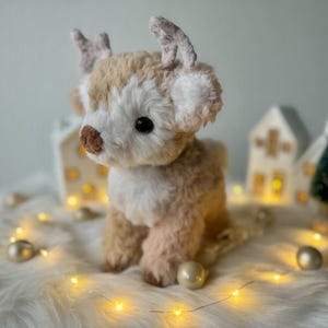 May include: A plush reindeer toy with a light brown and white fur coat, large black eyes, and small antlers. The reindeer is sitting on a white, fluffy surface, with decorative lights and miniature houses in the background.