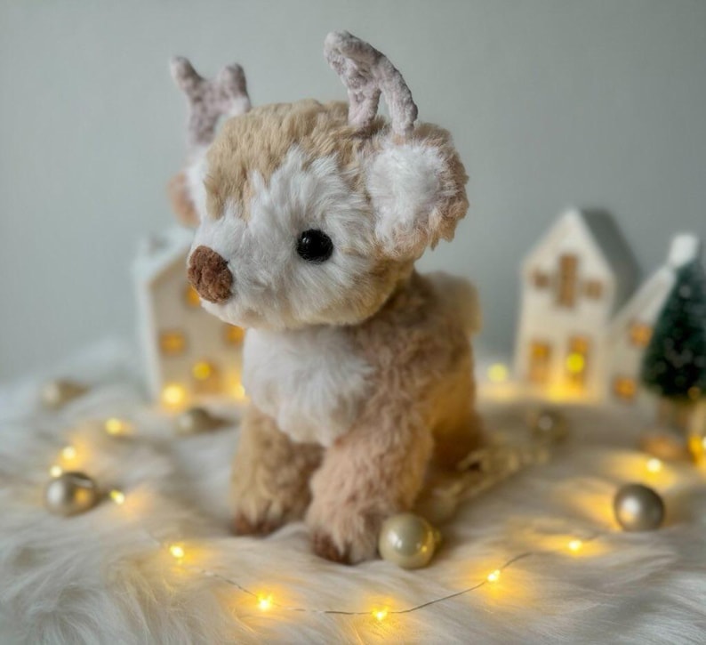 May include: A plush reindeer toy with a light brown and white fur coat, large black eyes, and small antlers. The reindeer is sitting on a white, fluffy surface, with decorative lights and miniature houses in the background.