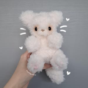 May include: A handmade, fluffy white teddy bear with large, dark eyes and pink paw pads. The plush toy has large ears and is held against a gray background. The artist's name, "pera.ftycookiee", is visible.