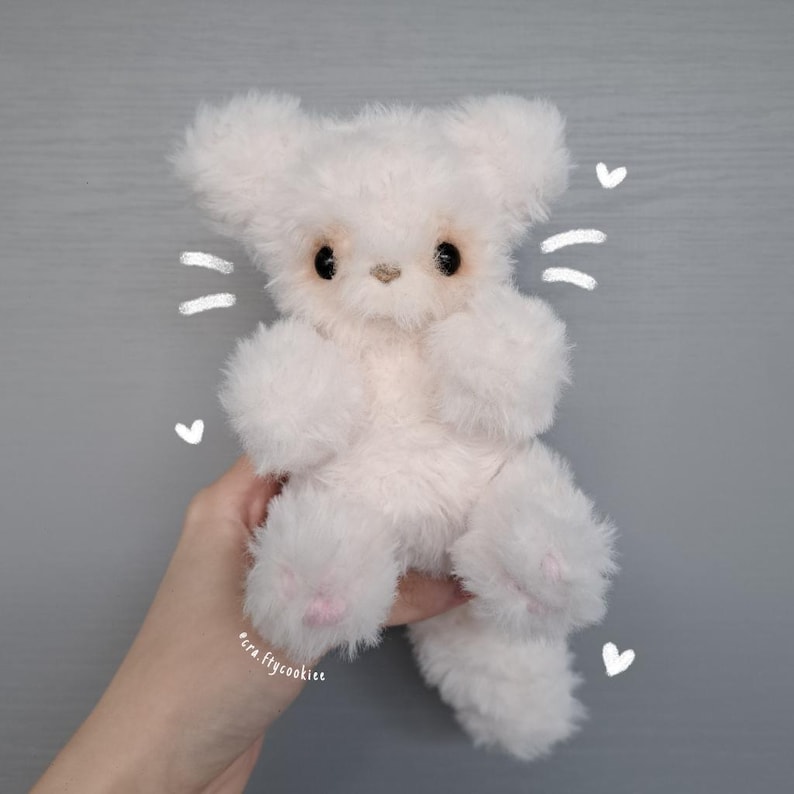 May include: A handmade, fluffy white teddy bear with large, dark eyes and pink paw pads. The plush toy has large ears and is held against a gray background. The artist's name, "pera.ftycookiee", is visible.