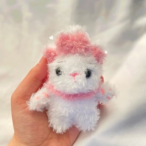 May include: A small, handmade white plush toy cat with pink accents. The cat has a pink hat, collar, and paw pads. It has black eyes and a pink nose. The toy is held in a person's hand.