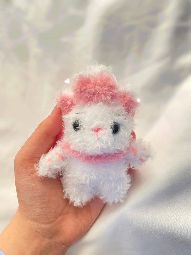 May include: A small, handmade white plush toy cat with pink accents. The cat has a pink hat, collar, and paw pads. It has black eyes and a pink nose. The toy is held in a person's hand.