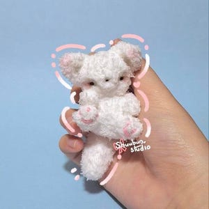 May include: A small, white plush toy in the shape of a bear, with pink accents on the ears and paws. The toy is held in a hand against a light blue background. The text "Shuubun studio" is visible.