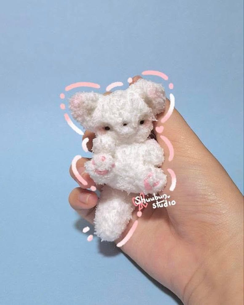 May include: A small, white plush toy in the shape of a bear, with pink accents on the ears and paws. The toy is held in a hand against a light blue background. The text "Shuubun studio" is visible.
