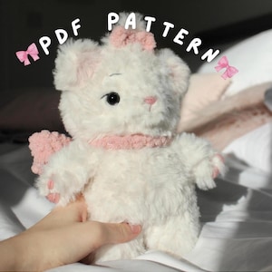 May include: A white plush cat toy with pink accents, including a bow, scarf, and paw pads. The text "PDF PATTERN" is displayed above the toy. The cat has black eyes and a pink nose.
