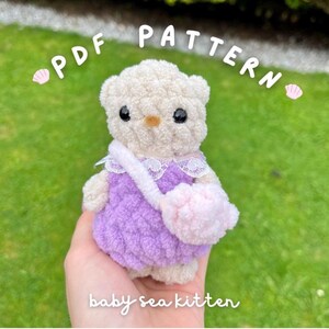 Op de afbeelding: Een handgemaakte, pluche babyzeekittenpop. De pop is crèmekleurig met zwarte ogen, een kleine oranje neus en een paarse jurk. Het heeft een roze tas en een witte riem. De tekst "PDF PATTERN" en "baby sea kitten" zijn zichtbaar.
