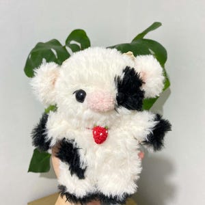 Pu&ograve; includere: Un peluche a forma di mucca con un motivo bianco e nero, con un naso rosa e un ciondolo a forma di fragola rossa. Il giocattolo ha piccoli occhi neri e orecchie soffici, progettato per essere morbido e coccoloso.
