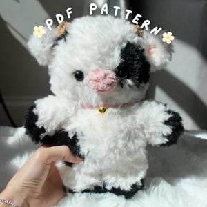 Puede incluir: Un peluche de vaca hecho a mano con un cuerpo blanco y esponjoso, manchas negras y una nariz rosa. La vaca tiene un collar rosa con una campana dorada. El texto "PDF PATTERN" está escrito encima de la vaca.