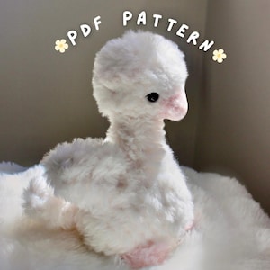 Puede incluir: Un cisne de peluche blanco con pico y patas rosas. El cisne está hecho de un material suave y esponjoso y tiene una expresión dulce e inocente. El texto "PDF PATTERN" está arqueado sobre el cisne.
