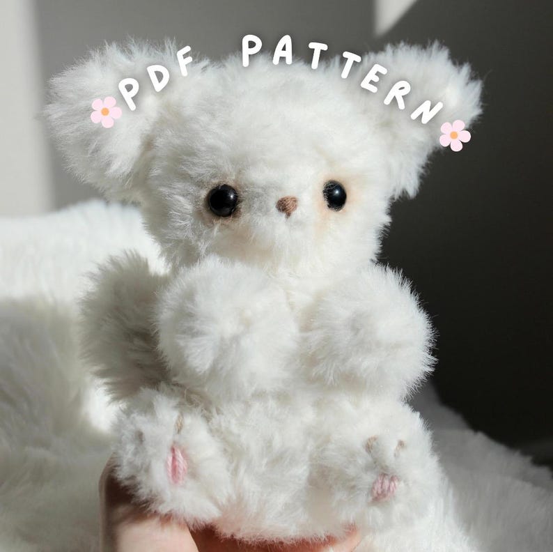 May include: A fluffy white teddy bear with black eyes and a small brown nose. The bear has pink accents on its paws and is labeled "PDF PATTERN" with flower accents. The bear is made of soft, plush material.
