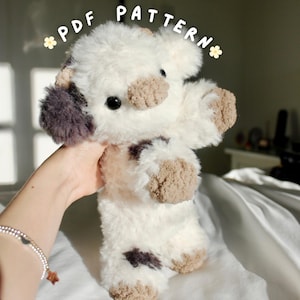 May include: A handmade plush toy in the shape of a cow, with a fluffy white body, brown and purple accents, and black eyes. The text "PDF PATTERN" is written above the toy. The toy is held by a person.