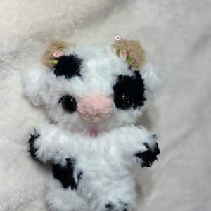 Pu&ograve; includere: Un giocattolo di mucca di peluche fatto a mano con un corpo bianco e soffice, macchie nere e un naso rosa. La mucca ha grandi occhi neri, piccole orecchie marroni con fiori rosa e zoccoli neri. Il giocattolo &egrave; progettato per assomigliare a un animale carino e coccoloso.
