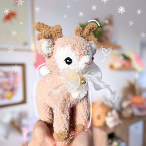 May include: A handmade plush reindeer with a cream-colored body, brown antlers, and a white bow with a small gold bell. The reindeer has black eyes and is held in a person's hands. The background has a blurred effect with snowflakes.