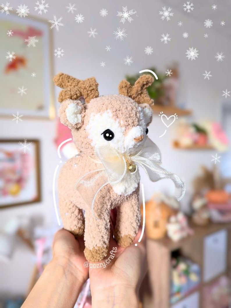 May include: A handmade plush reindeer with a cream-colored body, brown antlers, and a white bow with a small gold bell. The reindeer has black eyes and is held in a person's hands. The background has a blurred effect with snowflakes.