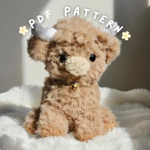 May include: A soft, brown Highland cow toy with white horns, black eyes, and a beige nose. It has a gold bell collar and the text "PDF PATTERN" is above the cow. The cow is sitting on a white, fluffy surface.