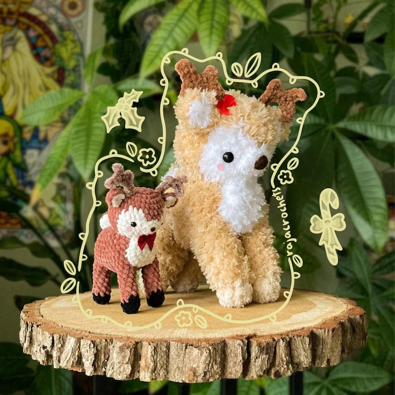 May include: Two crochet reindeer figurines, one large and one small, are displayed on a wooden slice. The larger reindeer is tan with white accents and a red bow. The smaller reindeer is brown with a red bow. The background includes greenery.