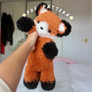 May include: A plush red panda toy with soft, textured rust-orange fur, black paws, black ear tips, and a white face patch. The toy features dark eyes and a small dark nose. The text "PDF PATTERN" is displayed in white letters above the toy.