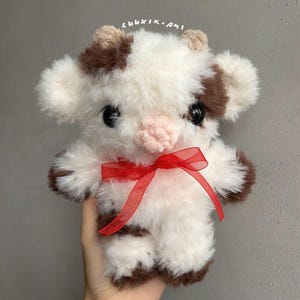 Pu&ograve; includere: Un giocattolo di mucca di peluche fatto a mano con un fiocco di nastro rosso. La mucca &egrave; bianca con macchie e accenti marroni, comprese le orecchie e gli zoccoli. Il giocattolo ha grandi occhi neri e un naso rosa. Il testo "luuxik.ami" &egrave; scritto in alto.