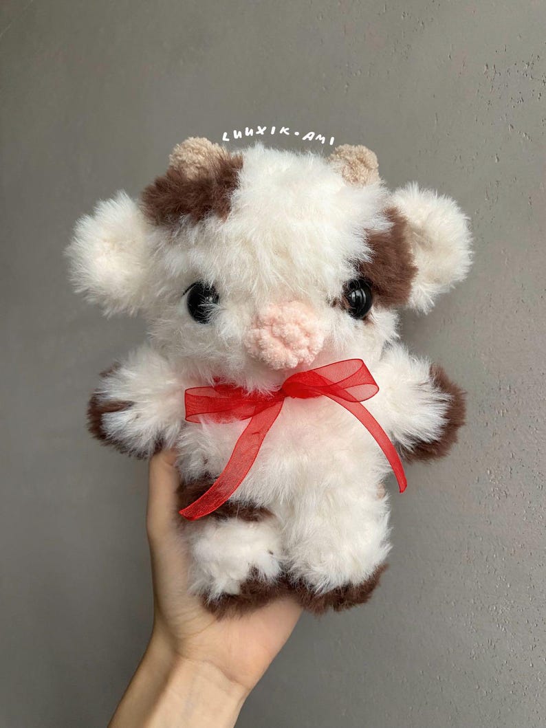 Pu&ograve; includere: Un giocattolo di mucca di peluche fatto a mano con un fiocco di nastro rosso. La mucca &egrave; bianca con macchie e accenti marroni, comprese le orecchie e gli zoccoli. Il giocattolo ha grandi occhi neri e un naso rosa. Il testo "luuxik.ami" &egrave; scritto in alto.
