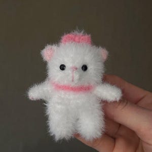 May include: A small, handmade white teddy bear with a pink bow on its head and a pink collar. The bear has black eyes and pink accents on its ears and paws. The plush toy is held in a hand.