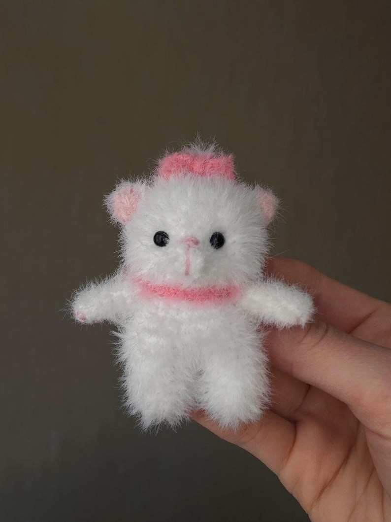 May include: A small, handmade white teddy bear with a pink bow on its head and a pink collar. The bear has black eyes and pink accents on its ears and paws. The plush toy is held in a hand.
